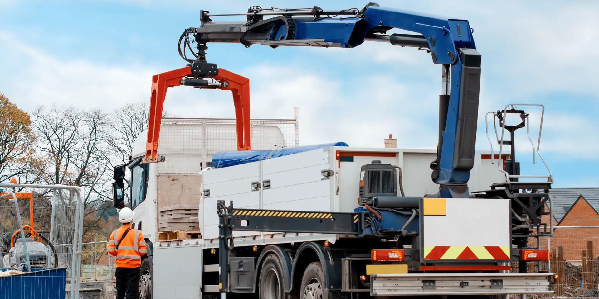 A lorry loader working on a construction site — knuckle-boom crane with a brick grab, stabilisers deployed, and a banksman in hi-vis and hard hat standing clear of the load