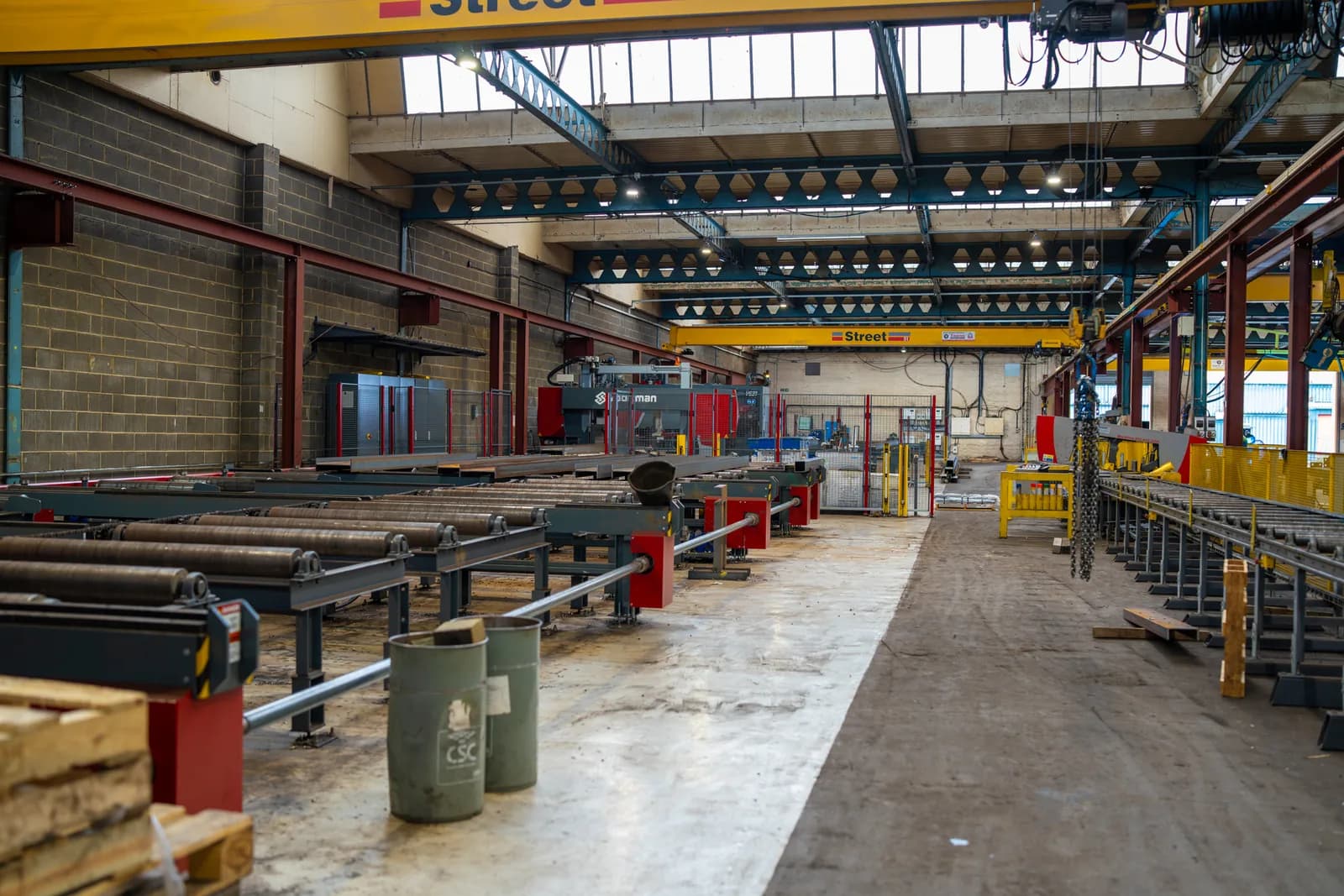 Yellow Street overhead travelling bridge cranes on runway rails above a roller-conveyor line in a UK steel-processing and fabrication workshop — fixed overhead lifting equipment that must be planned under LOLER 1998 and BS 7121-7