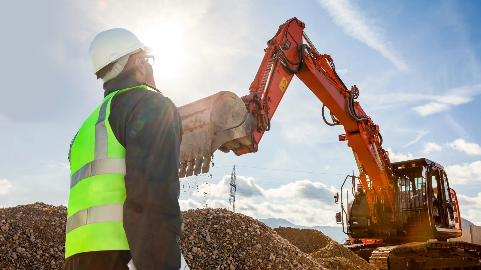 A construction supervisor in hi-vis and hard hat overseeing a 360° excavator on a UK construction site — an excavator lifting operation that must be planned and supervised under LOLER and BS 7121