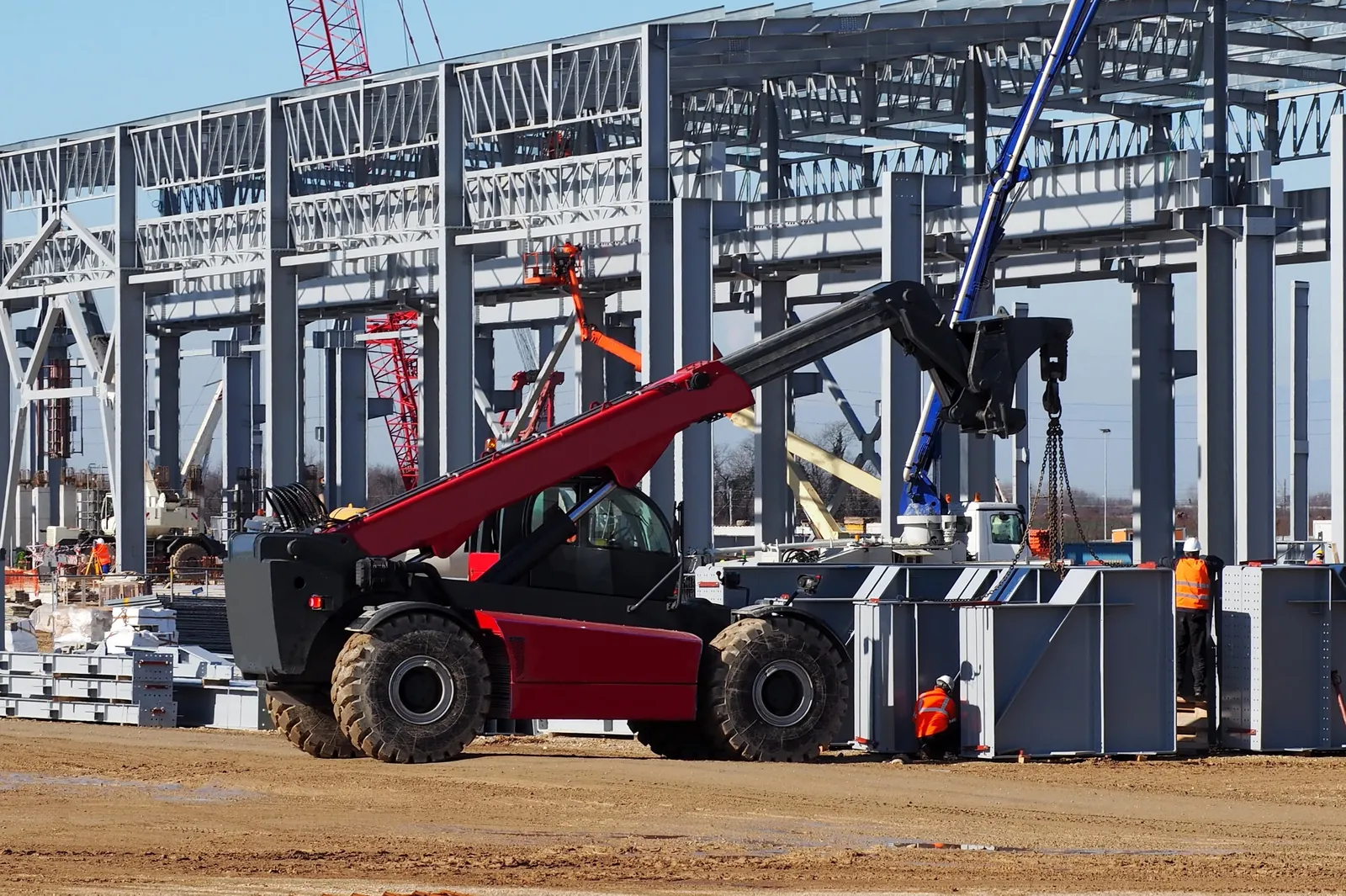 A red telehandler working alongside a mobile crane and banksman on a UK steel-frame construction site — the kind of suspended-load lifting operation that must be planned and supervised under LOLER and BS 7121